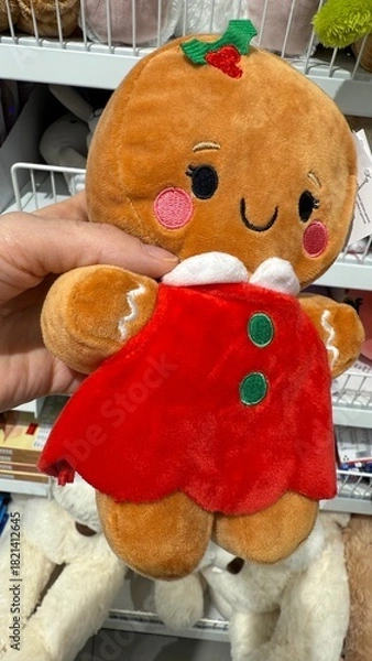 Fototapeta Adorable Plush Gingerbread Girl Toy in Red Dress with Holiday Details Held in Hand – Cute Christmas Stuffed Cookie Character on Store Shelf