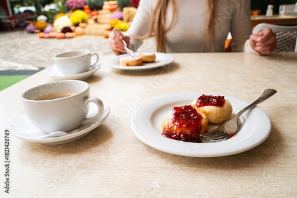Fototapeta Cottage cheese pancakes with jam served with coffee at cafe table, morning meal composition with female figure in soft background, warm food atmosphere
