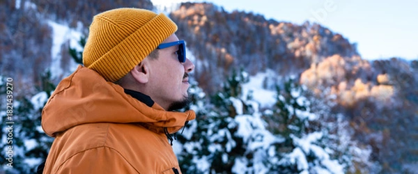 Fototapeta Man wearing orange jacket and yellow beanie against snowy mountains and trees , enjoying the tranquility of nature and beauty of outdoors in winter , copy space