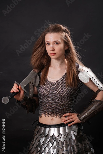 Fototapeta Portrait of a young woman, fantasy warrior in armor with a sword in her hands isolated on a dark background.