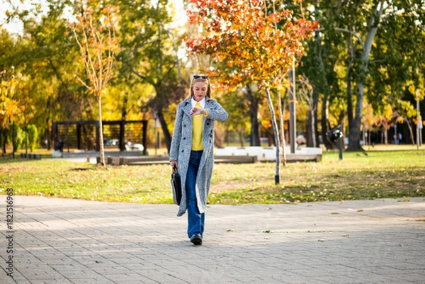 Obraz Confident businesswoman with sunglasses in stylish autumn coat checking the time on her wristwatch while walking with a laptop bag through the city park during sunny day.