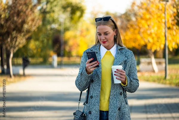 Obraz Portrait of urban businesswoman with sunglasses in modern autumn coat using smartphone and drinking coffee while standing in the city park during sunny day.