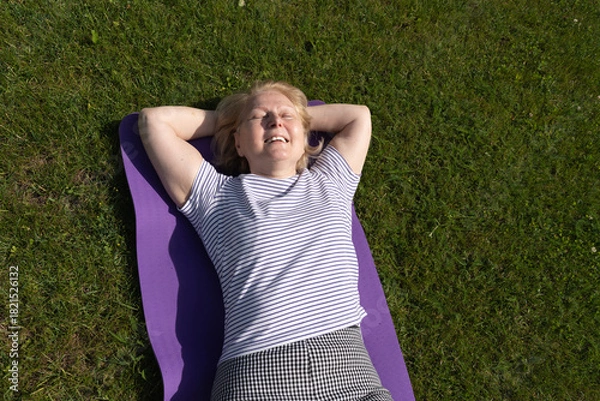 Obraz Senior woman relaxing on a yoga mat outdoors, lying on the grass with closed eyes and enjoying the sunlight. Peaceful moment of rest, wellness, and mindful outdoor lifestyle.