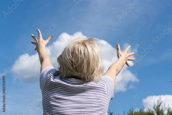 Obraz Senior woman raising arms toward the sky, enjoying a sunny day with blue sky and clouds. Seems like she is holding white cloud