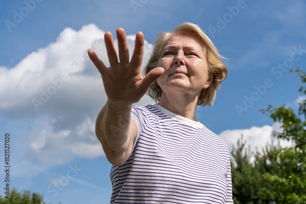 Obraz Senior woman reaching her hand forward toward the camera outdoors, standing under a blue sky with clouds. Strong expressive gesture conveying stop, attention, or protection.