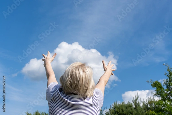 Obraz Senior woman raising arms toward the sky, enjoying a sunny day with blue sky and clouds. Seems like she is holding white cloud