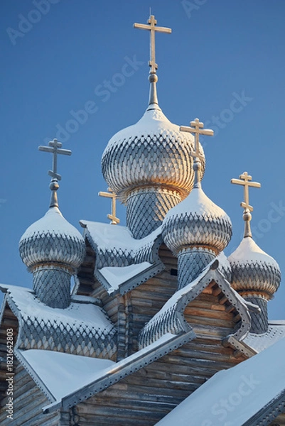 Fototapeta Wooden domes covered with snow and an Orthodox cross of the church against the blue sky in winter.