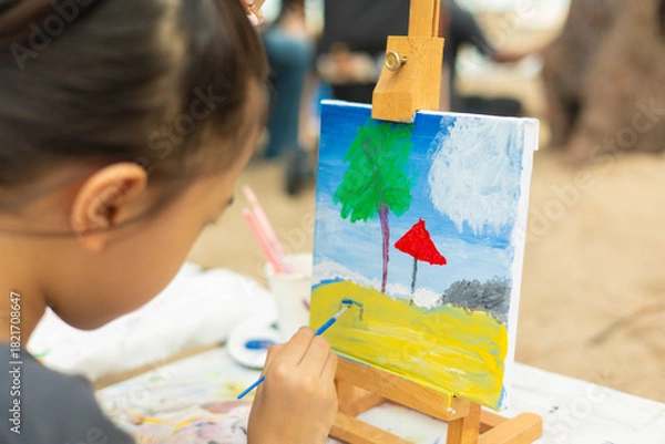 Fototapeta Close-up and selective focus of young girl painting a palm tree and sea with beautiful color on canvas. This shows creativity of a child's artistic hobby in a natural, outdoor beach setting.