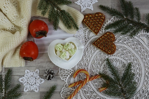 Fototapeta Christmas backdrop: a mug with sweets, persimmons, and cookies on a white lace napkin