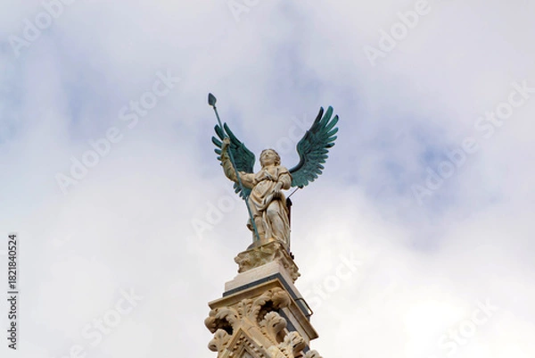 Obraz Angel Statue With Green Wings On Ornate Spire Against Cloudy Sky