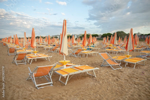 Fototapeta An empty sandy beach with rows of closed, colorful umbrellas and sun loungers in the warm evening light. The idea: preparation for the new beach season, a summer vacation, a quiet, uncrowded seaside r