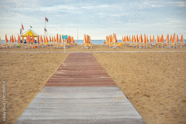 Fototapeta Wooden walkway leading to an empty sandy beach with rows of closed orange umbrellas and sun loungers, calm sea on the horizon, peaceful summer resort atmosphere.