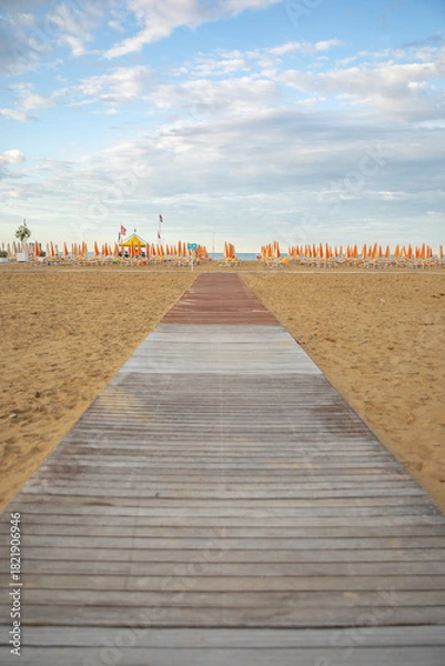 Fototapeta Wooden walkway leading to an empty sandy beach with rows of closed orange umbrellas and sun loungers, calm sea on the horizon, peaceful summer resort atmosphere.
