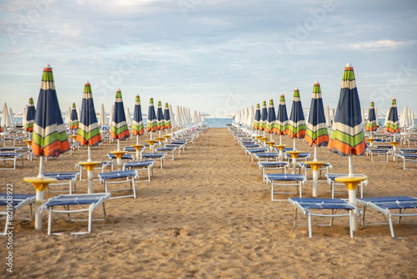 Fototapeta An empty sandy beach with rows of closed, colorful umbrellas and sun loungers in the warm evening light. The idea: preparation for the new beach season, a summer vacation, a quiet, uncrowded seaside r