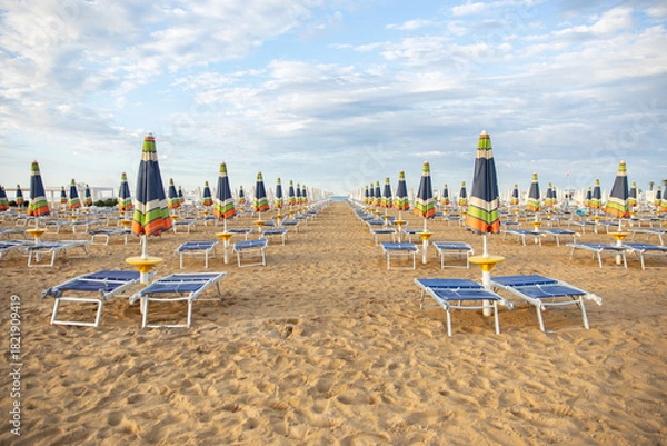 Fototapeta An empty sandy beach with rows of closed, colorful umbrellas and sun loungers in the warm evening light. The idea: preparation for the new beach season, a summer vacation, a quiet, uncrowded seaside r