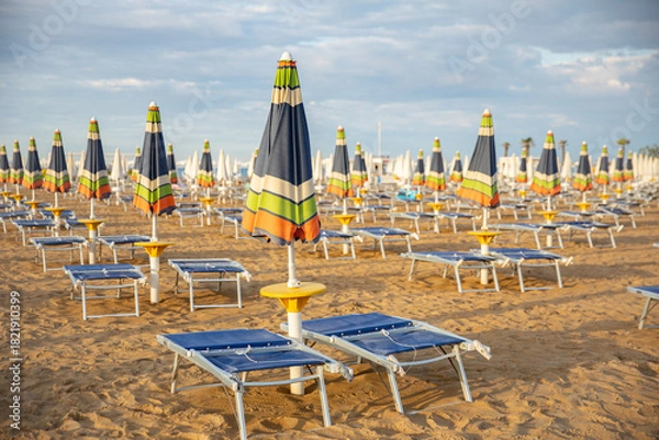 Fototapeta An empty sandy beach with rows of closed, colorful umbrellas and sun loungers in the warm evening light. The idea: preparation for the new beach season, a summer vacation, a quiet, uncrowded seaside r