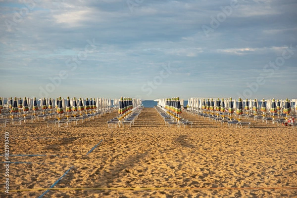 Fototapeta An empty sandy beach with rows of closed, colorful umbrellas and sun loungers in the warm evening light. The idea: preparation for the new beach season, a summer vacation, a quiet, uncrowded seaside r