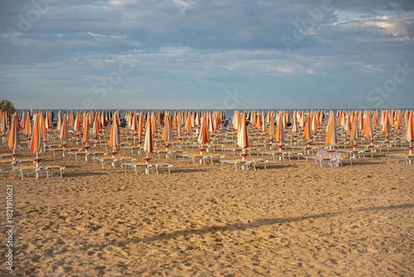 Fototapeta An empty sandy beach with rows of closed, colorful umbrellas and sun loungers in the warm evening light. The idea: preparation for the new beach season, a summer vacation, a quiet, uncrowded seaside r