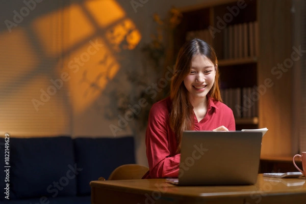 Fototapeta Young Asian woman smiling, doing remote work at home in the evening, using a laptop for online education