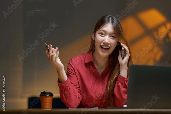 Fototapeta Happy young Asian professional laughing and talking on smartphone at desk, holding a pen while working remotely and planning tasks