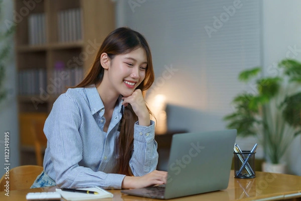 Fototapeta Happy Asian woman smiling, using a laptop, and working remotely from her home office, focusing on an online project