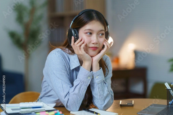 Fototapeta Young Asian woman wearing headphones, listening to music and daydreaming while taking a break from studying at her desk