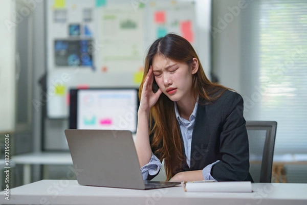 Obraz Stressed young Asian businesswoman suffering from severe headache or migraine while working on laptop in modern office, feeling burnout