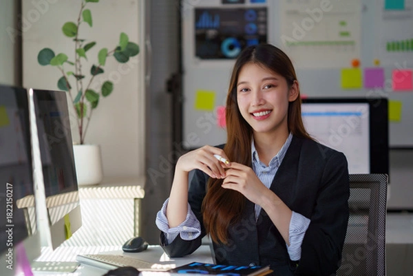 Fototapeta Young Asian businesswoman smiling while working with a computer at her desk, embracing success and professionalism in a startup office
