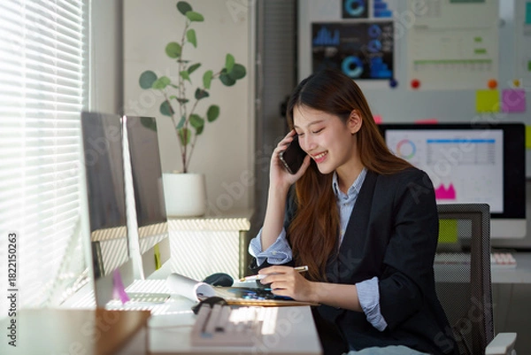 Fototapeta Smiling young Asian businesswoman managing her tasks, talking on the phone and writing notes while working at her office desk