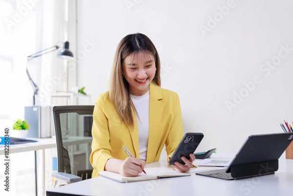 Fototapeta Young Asian woman smiling, writing notes in a notebook while holding a mobile phone and working at her modern desk