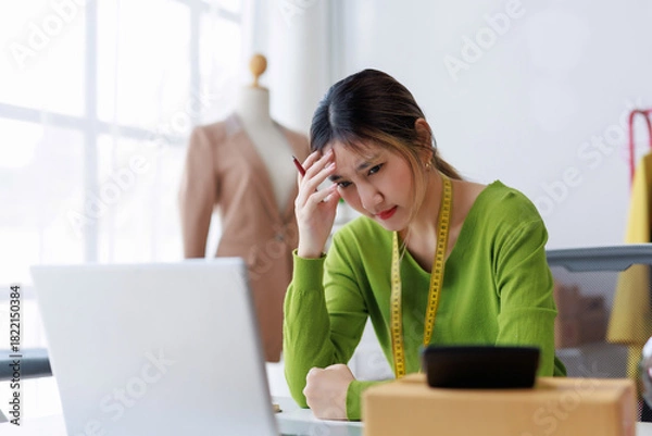 Fototapeta Stressed asian woman fashion designer having headache and looking at laptop while working in her small business studio