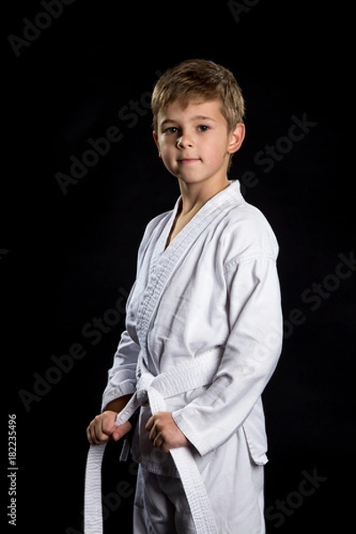 Obraz Smiling karate kid in brand new kimono, posing on the black background. Karate fighter holding his belt