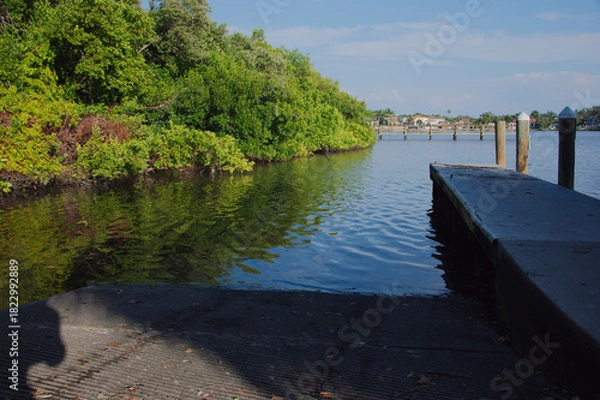 Obraz Tranquil Waterfront Dock Over Green bay With Reflective Water And Bright Sky Coffee Pot Park in St. Petersburg, FL. Wooden dock extending into clear water, lush green bushes along the bank, and a brig