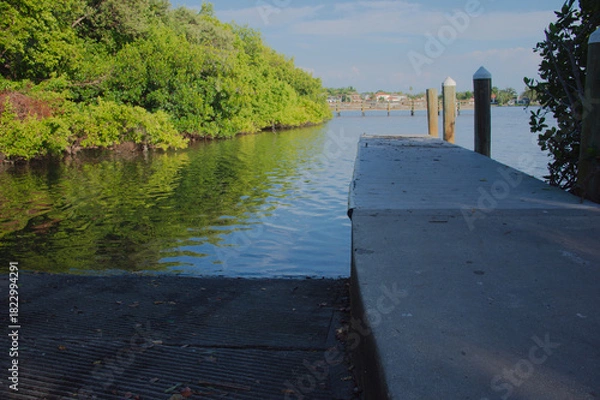 Obraz Tranquil Waterfront Dock Over Green bay With Reflective Water And Bright Sky Coffee Pot Park in St. Petersburg, FL. Wooden dock extending into clear water, lush green bushes along the bank, and a brig