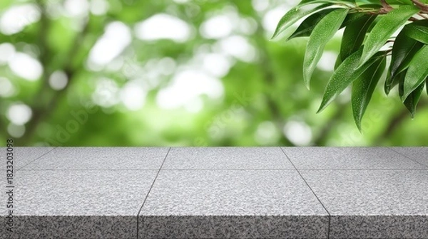 Fototapeta A clean, rough-textured gray granite stone tabletop or counter is positioned against a vibrant, defocused green forest background with natural bokeh, framed by dark green leaves sp