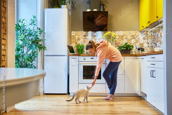 Obraz Happy woman stroking her charming cat while standing in her kitchen
