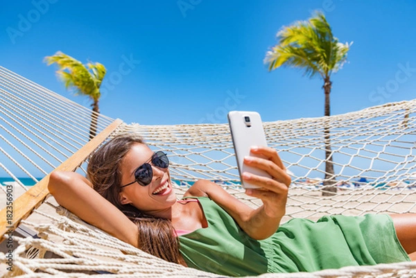 Obraz Letnia plaża styl życia młoda kobieta za pomocą SMS-ów aplikacji telefonu na smartfonie relaks w tropikalnym kurorcie karaibskim na hamaku. Mobilny internet Wi-Fi na wakacje w podróży Wakacyjna zabawa.