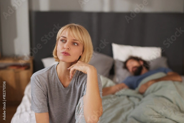 Fototapeta A woman appears thoughtful and concerned while seated on the edge of a bed. Her partner is asleep nearby, signaling tension and relationship problems.