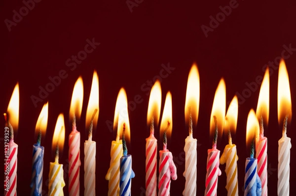 Fototapeta Row of colorful burning birthday candles on red background. Bright flames and vibrant colors with space for text or graphic elements. Ideal for celebration concepts.