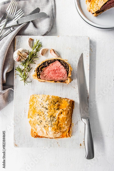 Fototapeta Homemade beef Wellington wrapped in golden puff pastry, sliced to reveal medium-rare beef tenderloin with mushroom duxelles. Styled on a rustic white background with garlic, herbs, and kitchen utensil