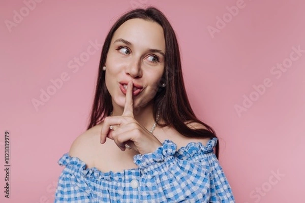 Fototapeta Woman in blue checkered top making a shushing gesture against a pink background while smiling at the camera