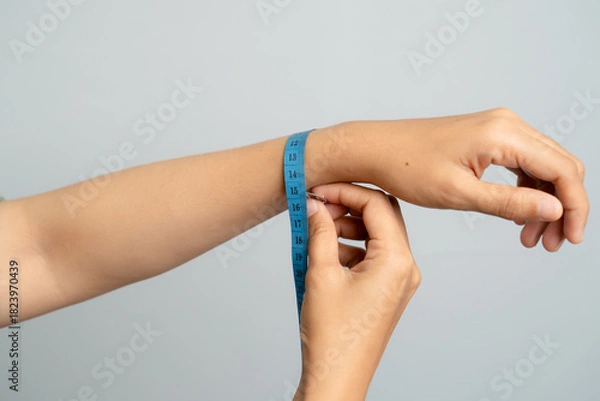 Fototapeta Close Up of a Woman Measuring Her Wrist Circumference with a Blue Tape Measure against a Soft Gray Background