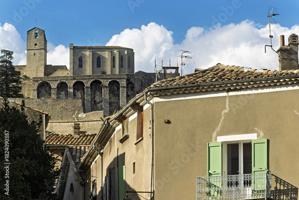 Fototapeta Traditional village scene in Sisteron, France, featuring beige stucco buildings and tiled roofs. A historic stone church with arched windows and a bell tower rises above the town
