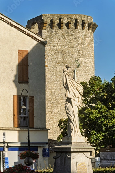 Obraz Stone statue of a robed figure holding an object aloft in a public square in Sisteron, France. Behind the statue stands a tall cylindrical tower with battlements, evoking medieval architecture.