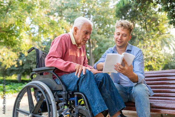 Fototapeta Grandson reading a book to his grandfather in a wheelchair while sitting on a bench at the park