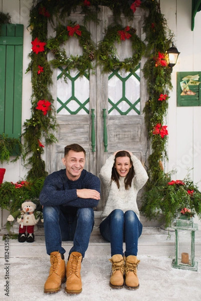 Fototapeta Happy funny young couple in love dressed in sweater sitting on porch steps at light house with decorated in red green New Year door at home. Christmas good mood. Family and holiday 2018 concept