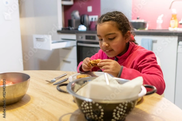 Obraz Young girl enjoying a delicious fried chicken dinner in a modern kitchen, focusing on the concepts of childhood, eating, and family meals at home