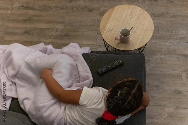 Fototapeta Young girl relaxing and watching tv on a comfortable couch at home, enjoying leisure time and entertainment while being cozy under a blanket