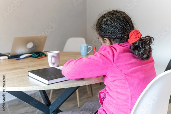 Obraz Young girl with braided hair and pink jacket busy concentrating on her schoolwork at a wooden table, diligently writing in her notebook during a study session