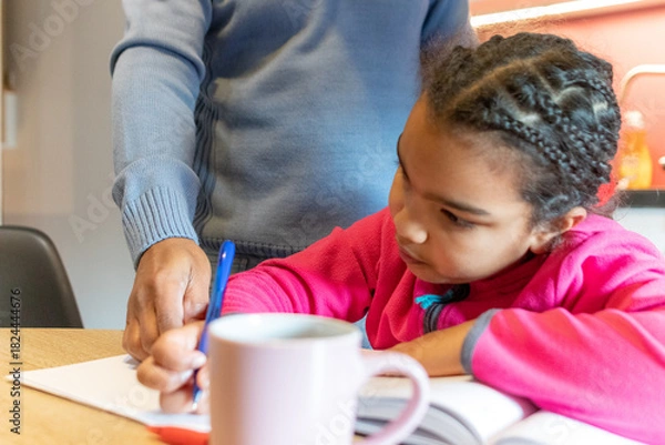 Obraz Little girl writing in a notebook, concentrating on her homework, with an adult male's hand gently guiding her in learning and education